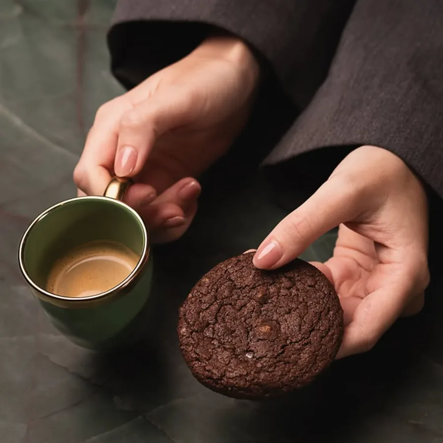 Mão segurando um cookie de chocolate ao lado de uma xícara de café.